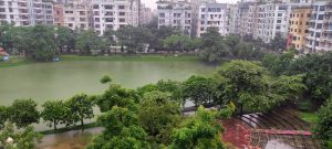Urban pond surrounded by multi-story residential buildings, green trees, and a paved park area with pink umbrellas under an overcast sky