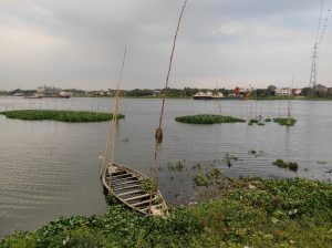 A weathered wooden boat rests by a riverbank under a cloudy sky. Lush greenery and floating plants dot the calm water, with buildings visible in the distance.