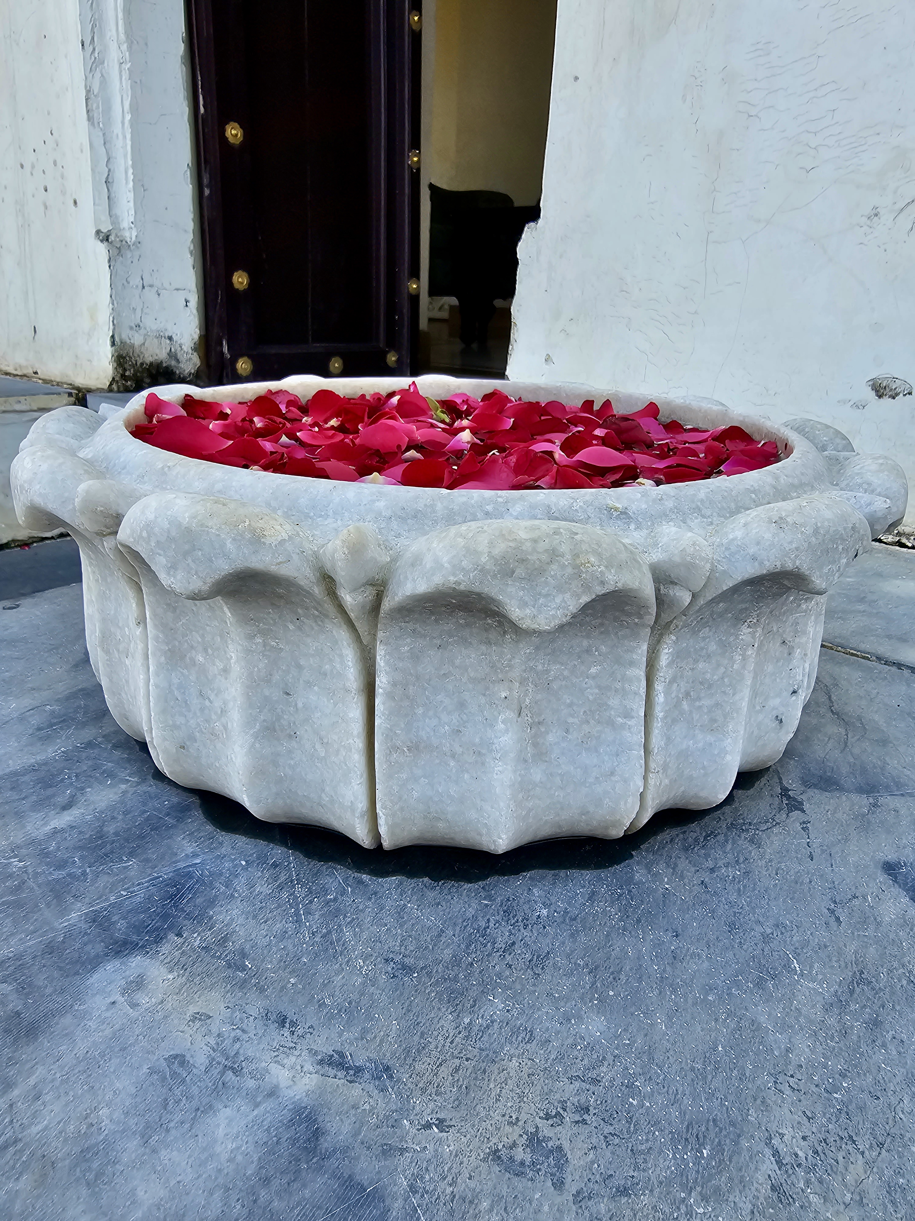 A white marble bowl filled with red rose petals is placed on the stone floor at Monsoon Palace, Sajjangarh, Udaipur. This bowl is used as a simple floral decoration near the palace entrance.