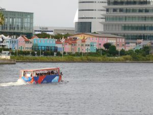 A colorful water bus travels on a river in the foreground, featuring passengers on board. In the background, a row of pastel-colored buildings with a variety of architectural styles, including windows and decorative elements, line the riverbank. 