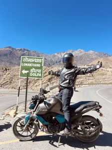 A person in a helmet and black leather jacket poses on a motorcycle, with a sign to Lomangthang and Dolpa against a mountainous blue-sky backdrop.
