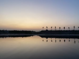 The photo captures a serene view of the Balu River in Bangladesh at dusk. The sky transitions from warm orange near the horizon to cool blue above, reflected perfectly in the calm river water. A line of silhouetted trees and distant structures stand along the riverbank, mirrored beautifully in the still surface below.