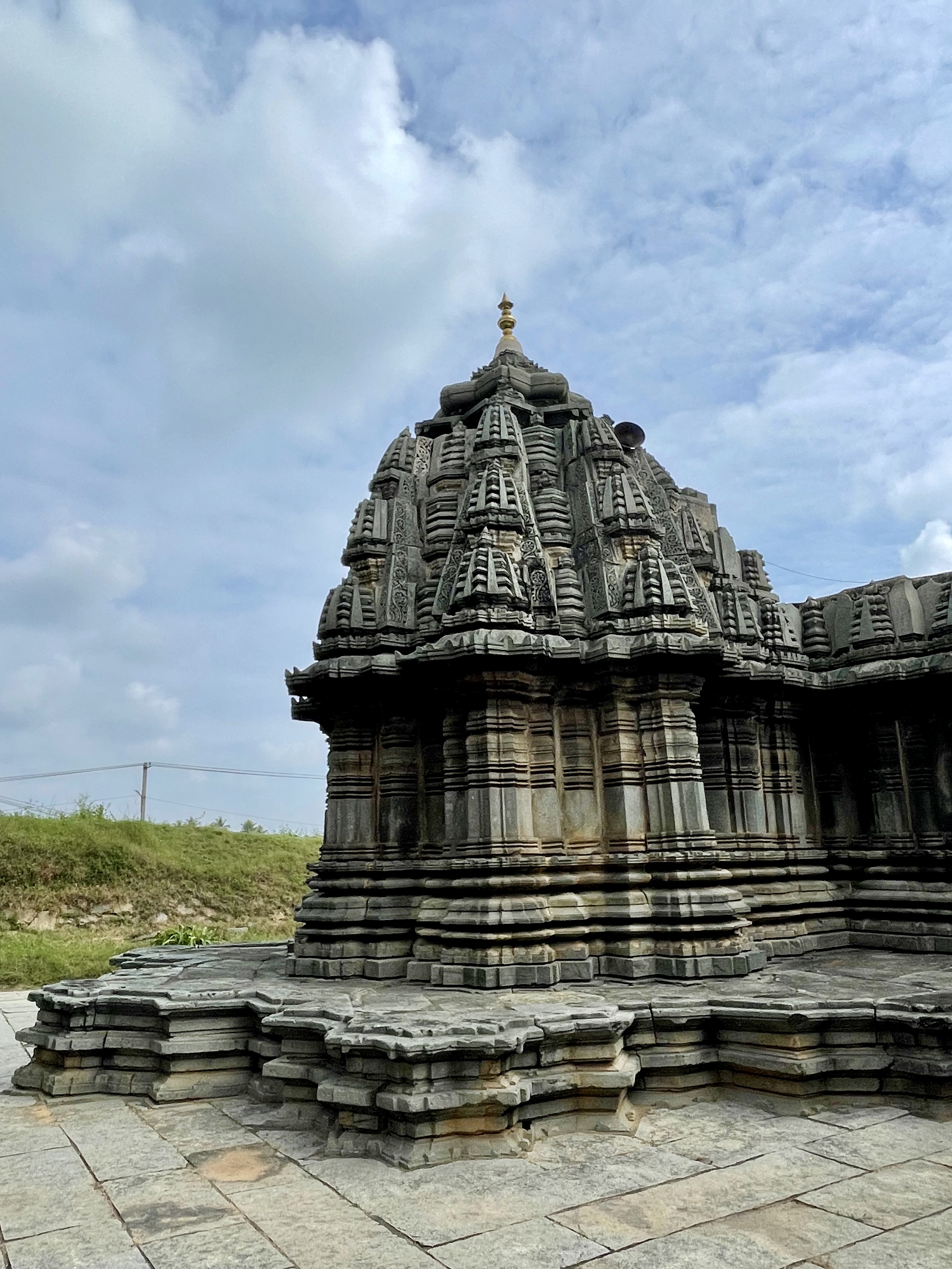 Close-up view of the shikhara (temple tower) at Lakshmi Narasimha Temple, Nuggehalli, Hassan. The detailed carvings and golden finial are captured under a dramatic, cloudy sky. 