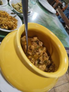 A yellow pot with chicken in reddish-brown sauce and a spoon inside, surrounded by small bowls of side dishes in a dining setting.