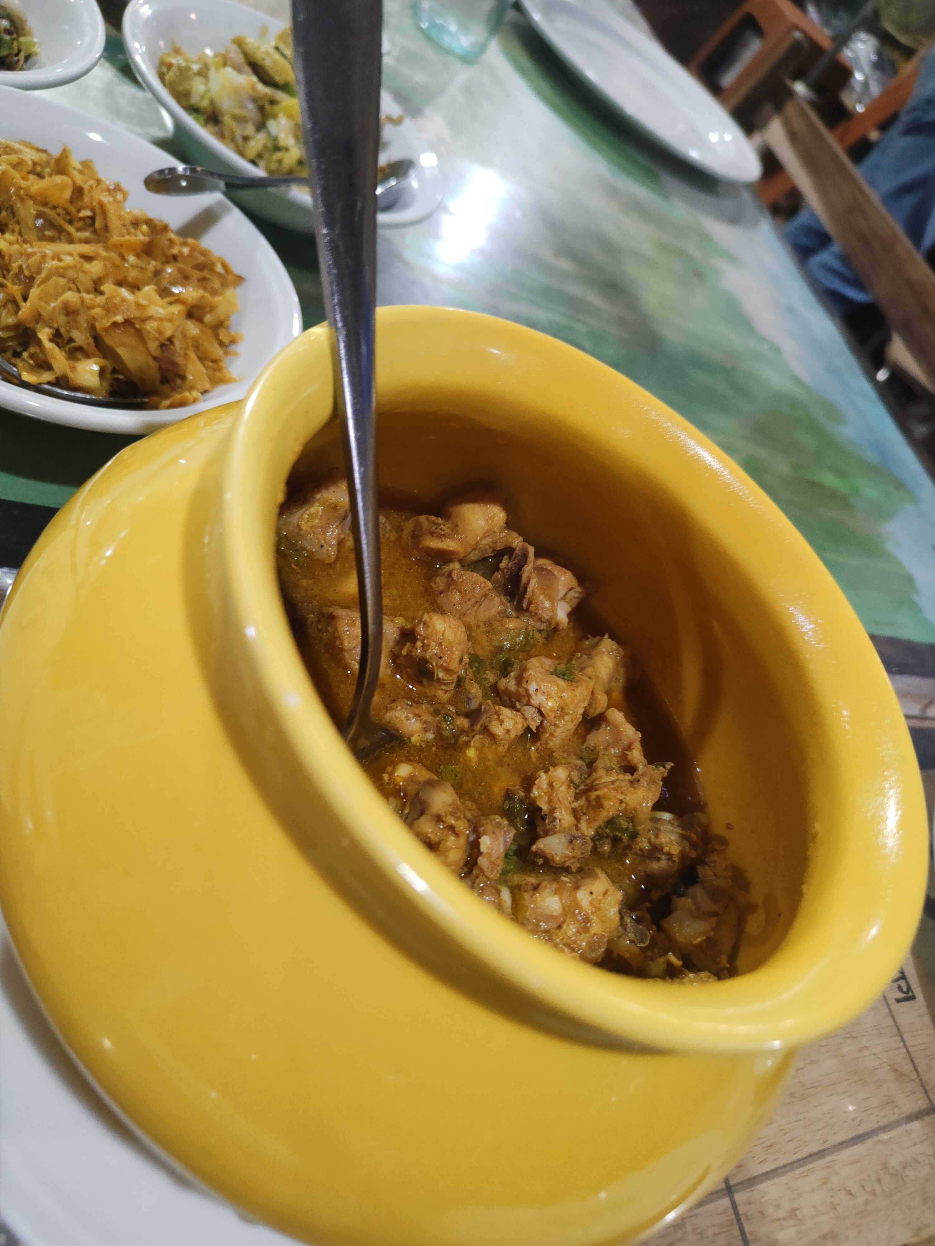 A yellow pot with chicken in reddish-brown sauce and a spoon inside, surrounded by small bowls of side dishes in a dining setting.