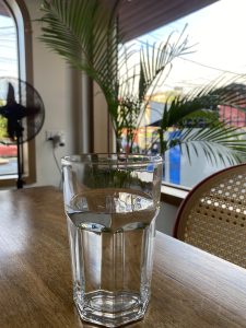 A glass of water on a wooden table with sunlight, a palm plant, and a fan in the background.