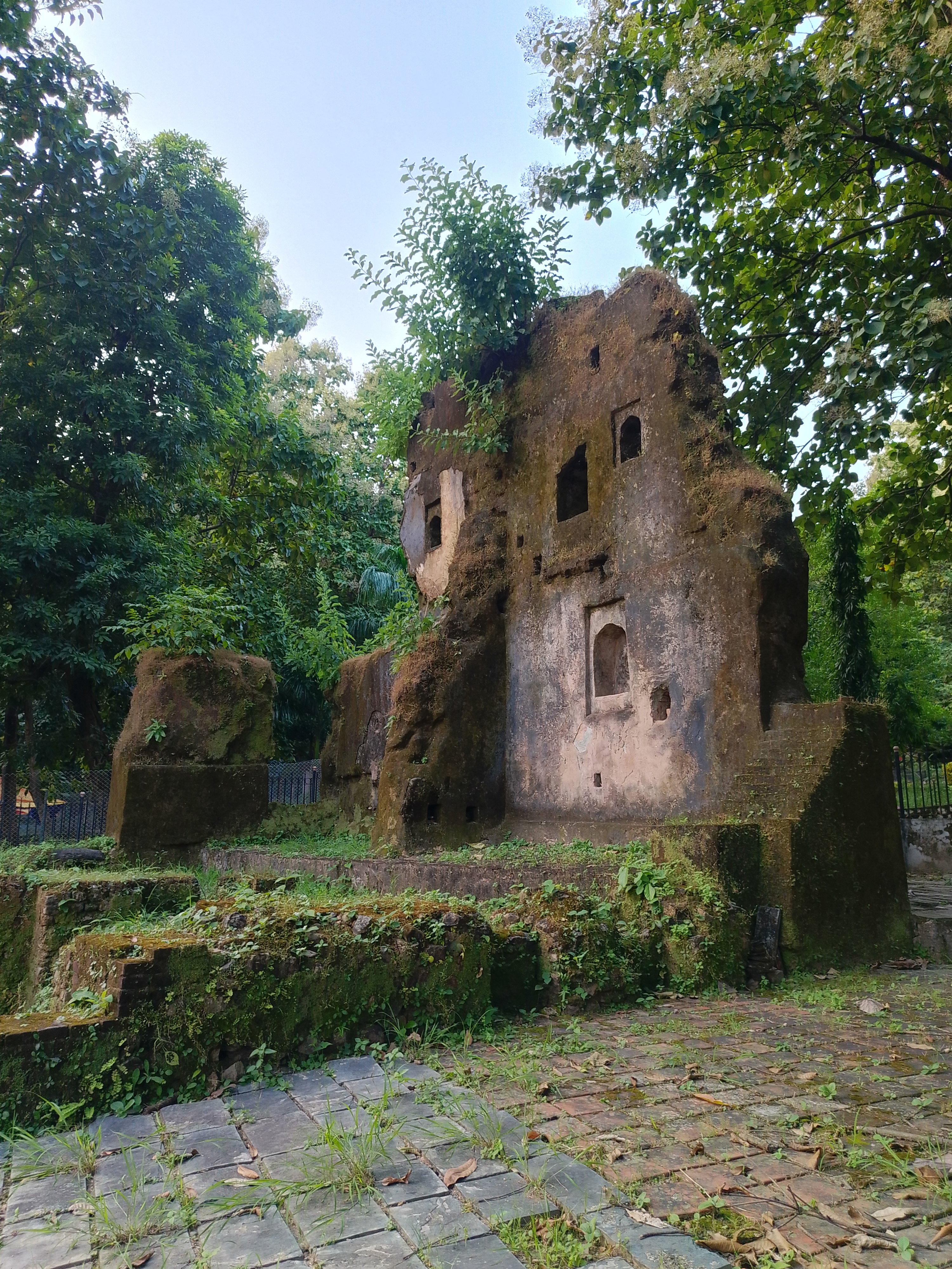 A partially collapsed stone structure, a ruin, is surrounded by lush green foliage.