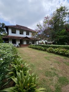 A wide view of a traditional building at Hill Palace, Thrippunithura, Kerala, with tiled roof, bright white walls, and a well maintained garden with flowering plants and trimmed grass. 