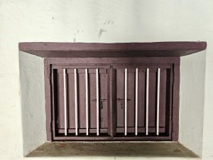 A tiny wooden window secured with vertical iron bars and a latch, set within a thick wall, seen at Hill Palace, Thrippunithura, Kerala. 