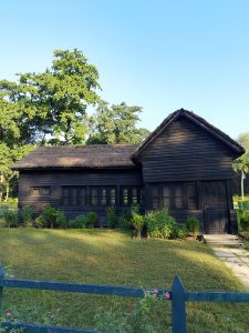 A small, dark wooden house with a thatched roof stands in a grassy yard surrounded by green plants and trees under a clear blue sky. Sunlight highlights parts of the roof and lawn, and a low fence with a few flowers runs along the foreground.