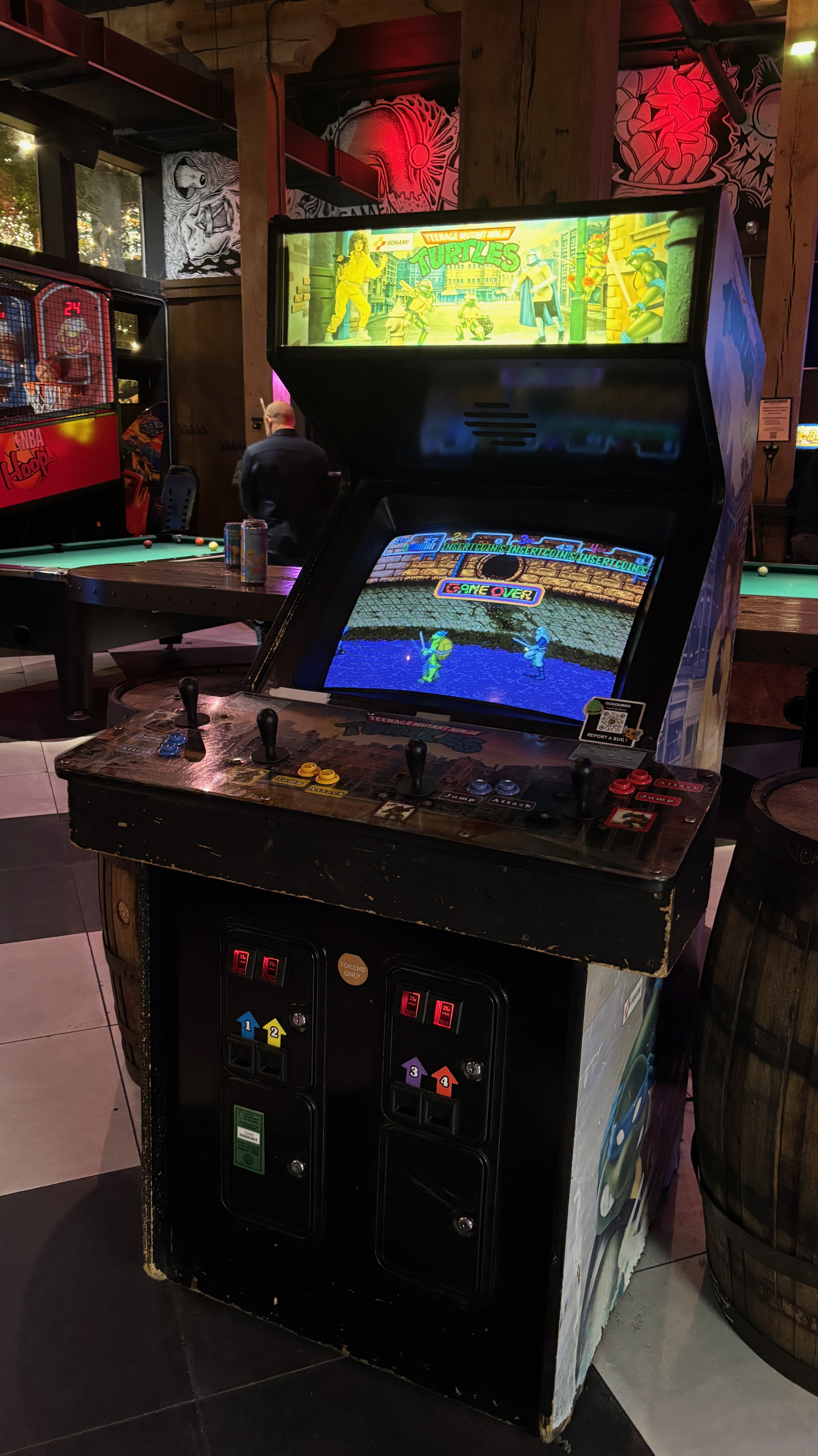 An old stand-up video arcade game from the 1980s. It appears in a bar setting with a few cans on the table. Its background features a pool table with one man who appears to be playing, holding a stick.