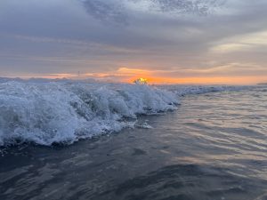 A close-up of a wave crashing in the ocean at sunset, with the sun&#039;s orange glow visible just above the water on the horizon and streaky clouds overhead