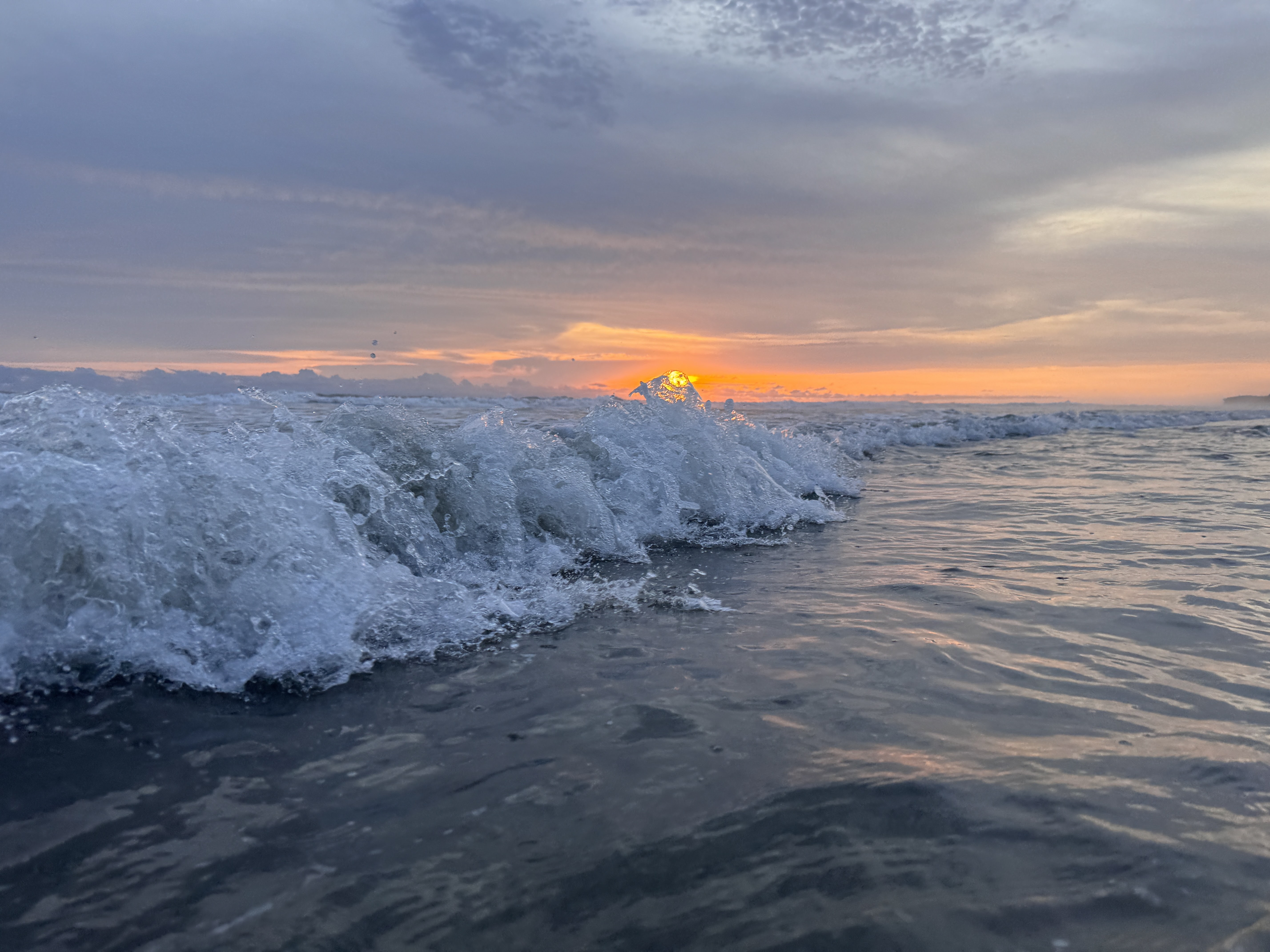 A close-up of a wave crashing in the ocean at sunset, with the sun's orange glow visible just above the water on the horizon and streaky clouds overhead