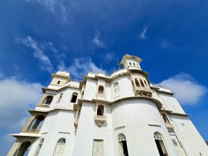 A wide-angle view of the white Monsoon Palace at Sajjangarh, Udaipur, standing tall under a clear blue sky. The palace is known for its royal architecture and hilltop location. 