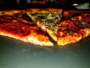 A close-up view of three pizza slices on a dark plate, featuring pepperoni, mixed meat with herbs, and a white-sauce slice with cheese, all with golden crispy crusts.
