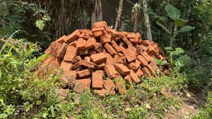 A pile of red bricks is stacked in a garden setting, surrounded by various green plants and foliage.