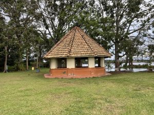 A small polygon shaped building with no windows stands with trees and a lake in the background.