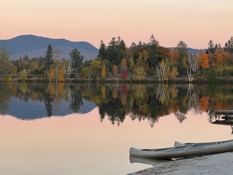 A calm lake at sunset reflects a forest of autumn-colored trees and distant mountains under a soft pink sky. Two silver canoes rest on the sandy shore in the foreground, while the still water mirrors the vibrant orange, yellow, and green foliage along the opposite bank.