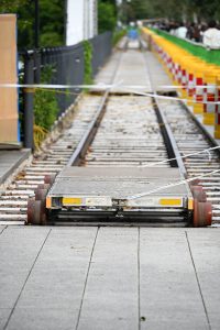 A close-up of a light rail track with a flatbed cart on the rails, surrounded by a paved walkway, bright construction barriers, scattered debris, and blurred people near a green fence.