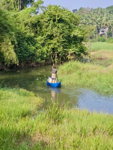 A man paddling a small fiber boat in a narrow canal surrounded by lush greenery. Taken in Ayamkulam, Mavoor, Kozhikode, reflecting peaceful village life. 