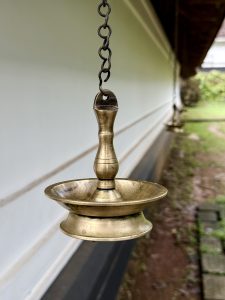 A close-up of a traditional brass hanging lamp (Nilavilakku) suspended from a chain, photographed against the white wall of a temple in Kozhikode. Captured in soft daylight, it highlights the cultural and spiritual essence of Kerala architecture.