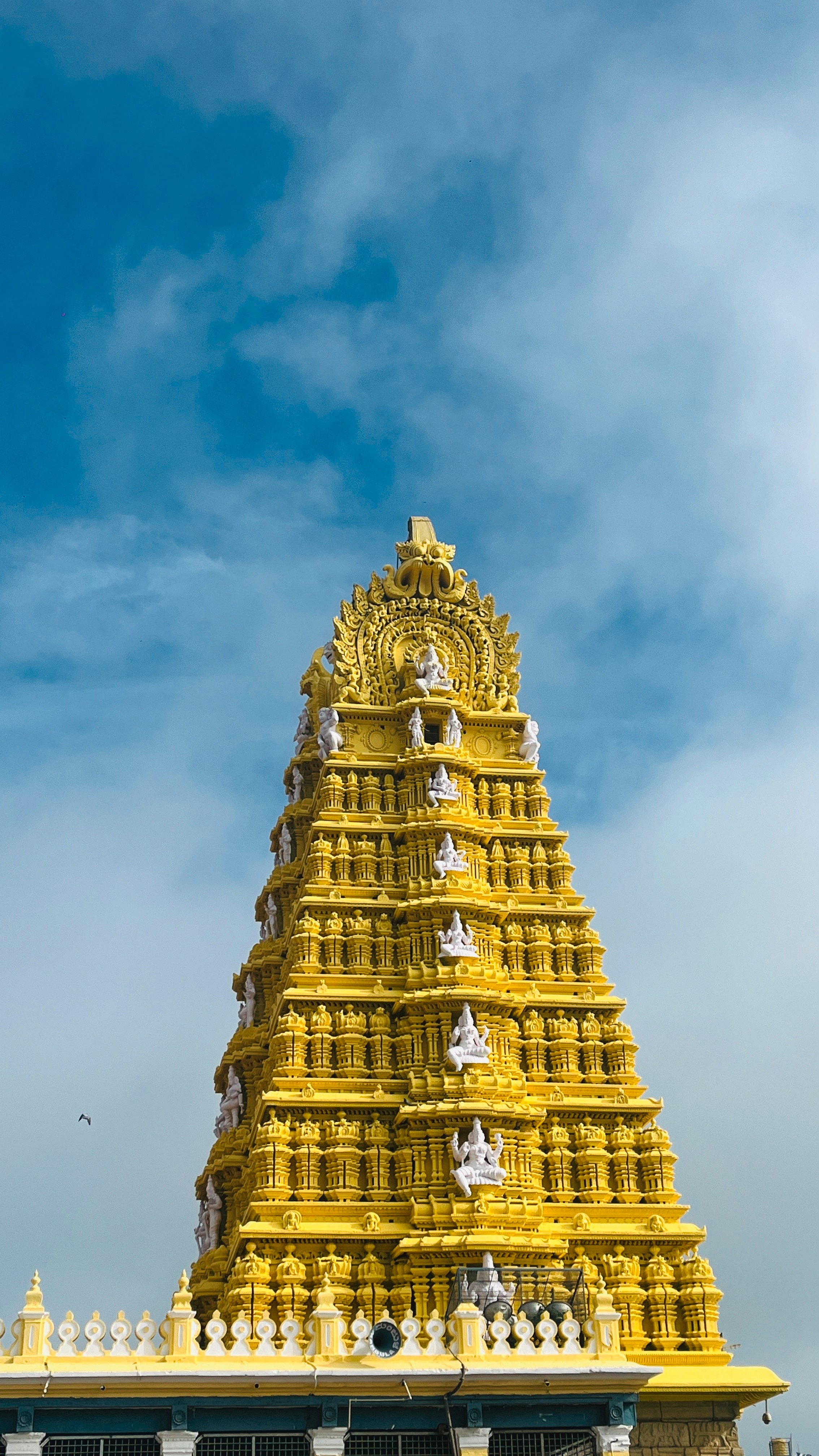 A tall, intricately designed chamundi temple painted in vibrant yellow, adorned with white sculptures and decorative patterns.