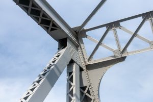Close-up view of a steel bridge structure showing intersecting beams, rivets, and metal joints against a light blue sky. The geometric framework emphasizes the industrial design and strength of the bridge.