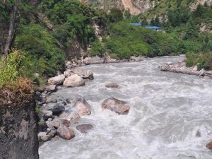 A rushing river flows through a rocky landscape, surrounded by lush green vegetation. Smooth stones and large boulders are visible along the water's edge, with steep, forested hillsides rising on both sides.