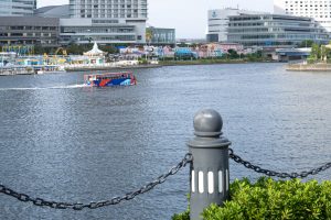 A colorful boat is cruising on a river, In the background, a row of vibrant, multi-colored buildings can be seen, along with a carousel and modern structures in the distance.