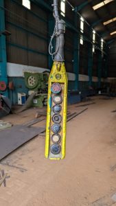 A close-up of a worn yellow industrial control panel with multiple buttons hanging inside a large warehouse. The background features machinery and industrial equipment.