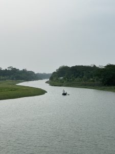A raw image showing a winding river on a cloudy day. The river curves between a flat, grassy bank on the left and dense, green trees on the right. A small, dark boat with two figures is visible in the center of the water