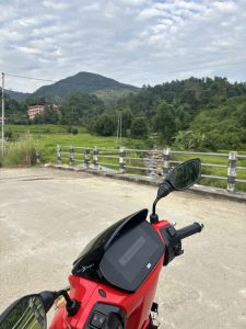 Red scooter on concrete with digital dashboard, against green hills and partly cloudy sky.
