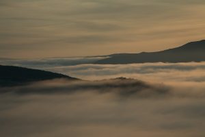 A tranquil landscape featuring rolling hills partially covered in soft, low-lying fog. The scene is bathed in warm hues of orange and gold, suggesting either sunrise or sunset, with a clear sky above and a silhouette of distant mountains on the horizon.