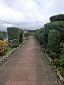 A stone path lined with manicured topiary trees leads through a lush garden under a cloudy sky. The scene is peaceful and neatly organized.