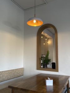 A cozy indoor scene with a wooden table, beige bench, warm light, and a mirror reflecting a plant.