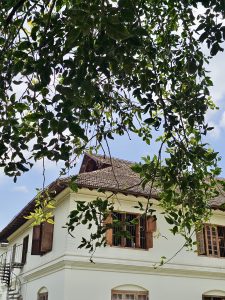 A bright corner view of a heritage building at Hill Palace, Thrippunithura, Kerala, with open wooden windows, tiled roof, and surrounding green tree branches in the foreground. 