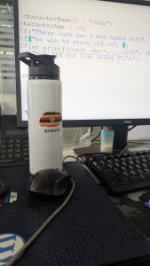 A white water bottle with the Ugandan flag and the word “MUKONO” is standing on a desk in front of a computer monitor that displays lines of code.
