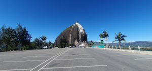 A large rock formation stands prominently against a clear blue sky, partially shaded by green foliage from palm and pine trees. In the foreground, an empty parking lot with several parking spaces is visible. In the background, the rock features a staircase leading up its side, and there are small buildings and attractions at its base. The landscape surrounding the rock shows distant hills, enhancing the scenic view.