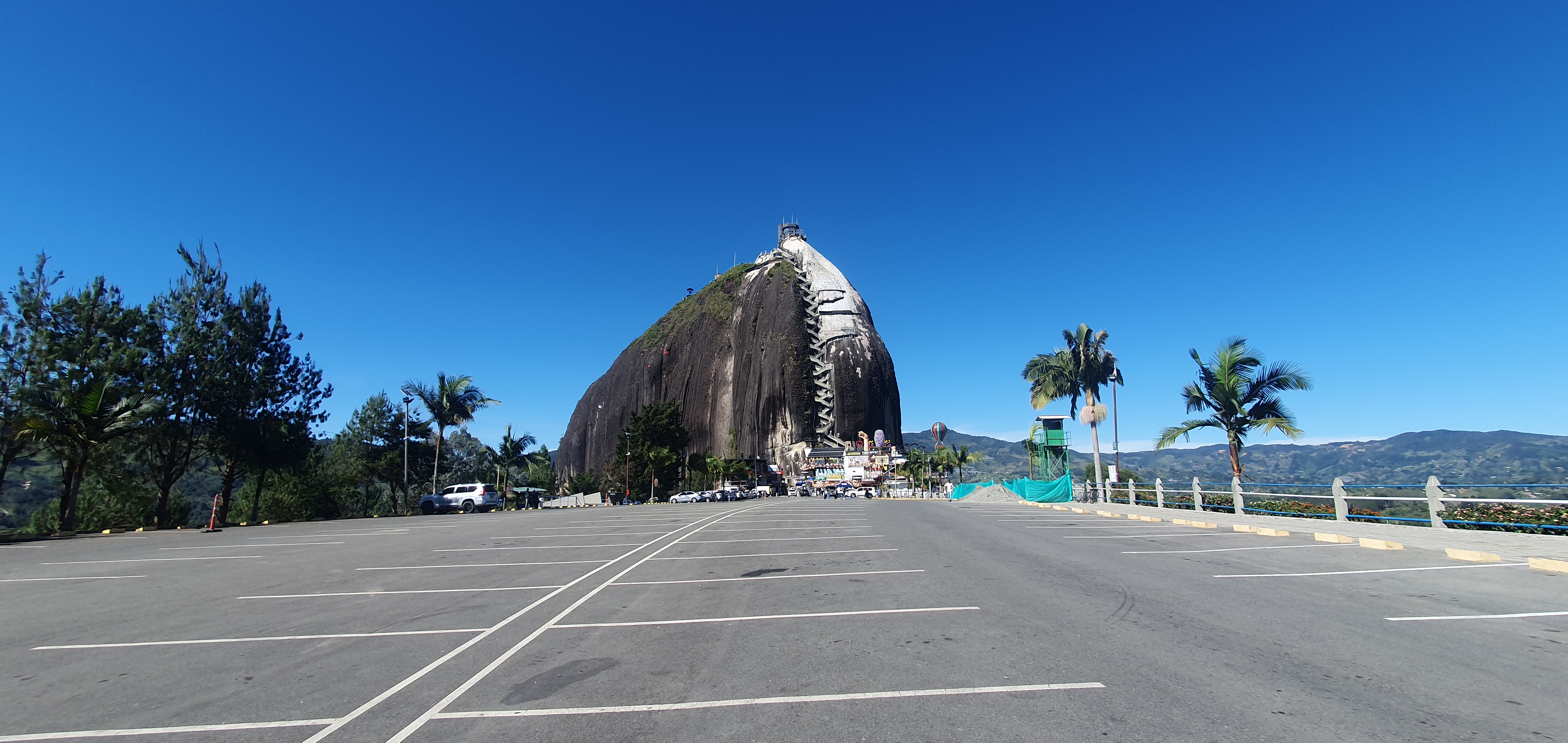 A large rock formation stands prominently against a clear blue sky, partially shaded by green foliage from palm and pine trees. In the foreground, an empty parking lot with several parking spaces is visible. In the background, the rock features a staircase leading up its side, and there are small buildings and attractions at its base. The landscape surrounding the rock shows distant hills, enhancing the scenic view.