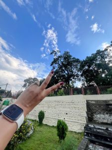 A person&#039;s hand wearing a white smartwatch and a ring flashes a peace sign against a clear blue and cloudy sky. Below the hand is a white textured retaining wall and grass.