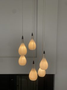 Six teardrop pendant lamps with frosted glass hang from thin cords, glowing warmly against a white wall.