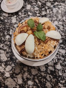 A close-up view of a bowl of biryani, featuring saffron-colored rice mixed with caramelized onions, cashews, and halved boiled eggs, garnished with fresh mint leaves