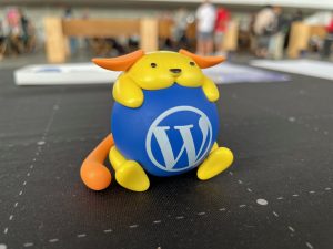 A small yellow Wapuu mascot hugging a blue ball with the white WordPress &ldquo;W&rdquo; logo, sitting on a black tabletop with a blurred event crowd in the background.
