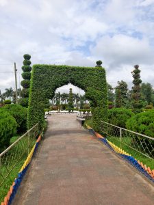 A pathway leads through an archway covered in lush green foliage, flanked by neatly trimmed hedges and ornamental trees. 