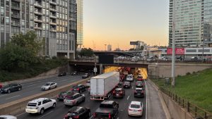 A downtown freeway, crowded with cars. In the background there are tall buildings on either side of the freeway, with a bit of evening sky showing in the middle. Chicago.