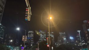 A city skyline at night. In the top left, quite close, is a stop light pointed to the left.  In the center is a bright street light with strong lens flare around it.