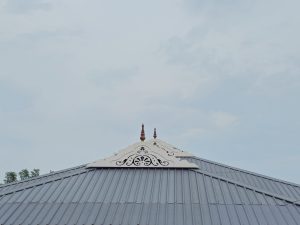A grey sloped roof with traditional white and wooden decorative ornaments at the top, under a clear blue sky. Taken in rural Kozhikode, Kerala. 