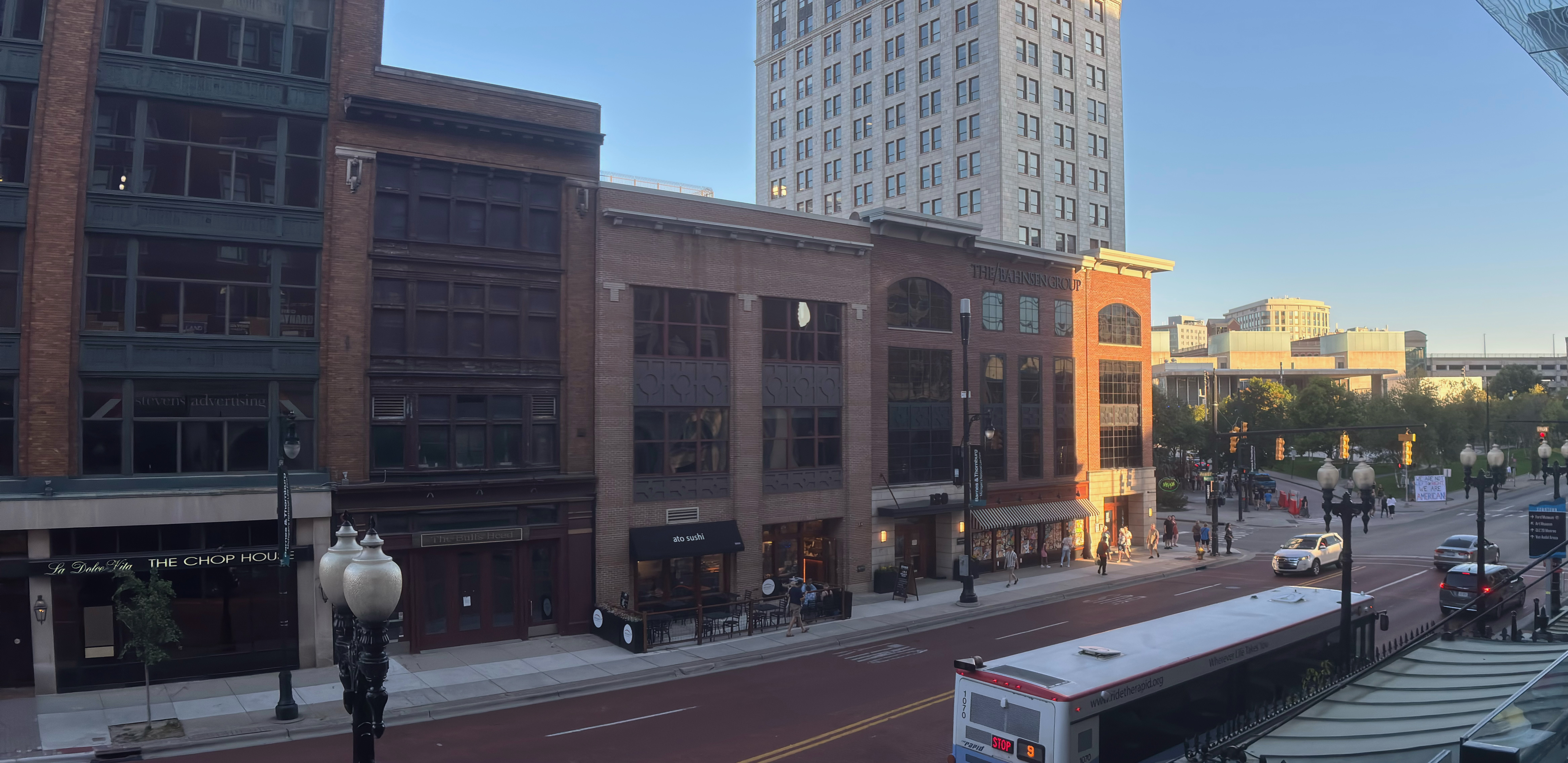 A city street scene. The sun is low, so some buildings are lit, whilst some or shadowed. Cars, busses, and people are on the street. Grand Rapids, Michigan.