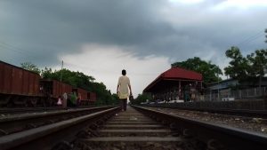 A Bangladeshi boy walks barefoot on the railway, shoes in hand, lost in thought as the storm clouds gather. The rails stretch ahead — uncertain, yet familiar — just like his dreams.
