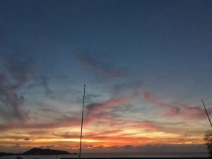 A scenic ocean sunset with a colorful sky of orange, pink, and blue hues, wispy clouds, and a distant hill silhouette, framed by thin poles in the foreground.